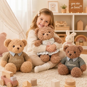 Könnte beinhalten: Drei Plüschbären mit gestickten Namen. Ein Bär trägt einen beigen Pullover mit der Stickerei 'Oliver', einer ein blau-weißes Blumenkleid mit der Stickerei 'Lily' und einer einen salbeigrünen Pullover mit der Stickerei 'Thomas'.