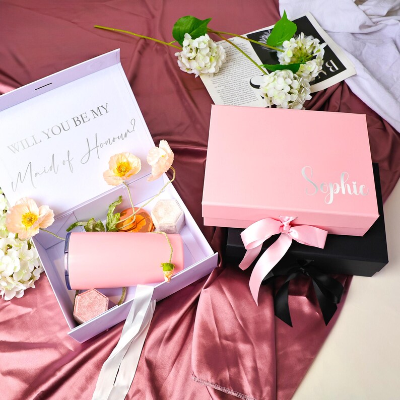 May include: Two pink gift boxes, one open and one closed, with floral accents. The open box contains a pink tumbler, a small hexagonal box, and other items. The closed box has the name "Sophie" in silver script. The text "Will you be my Maid of Honour?" is visible.