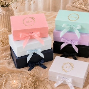 Pode incluir: Uma coleção de caixas de presente coloridas, cada uma com uma fita de cetim combinando. As caixas são em tons de rosa, verde menta, lavanda, preto, branco e azul marinho. Algumas caixas têm letras douradas e desenhos florais.