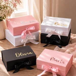 May include: A collection of gift boxes in various colors, including pink, white, and black, each adorned with a name in gold or silver lettering. The boxes are tied with satin ribbons in coordinating colors, creating an elegant presentation. The names on the boxes are: Kasia, Olivia, and Marta.