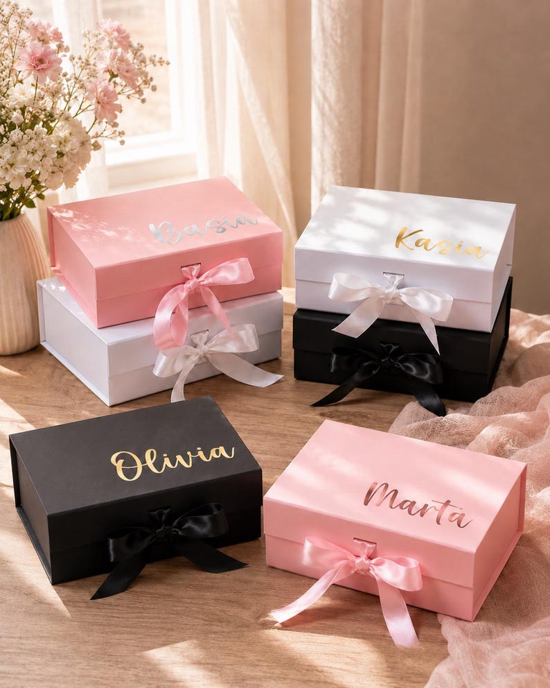 May include: A collection of gift boxes in various colors, including pink, white, and black, each adorned with a name in gold or silver lettering. The boxes are tied with satin ribbons in coordinating colors, creating an elegant presentation. The names on the boxes are: Kasia, Olivia, and Marta.