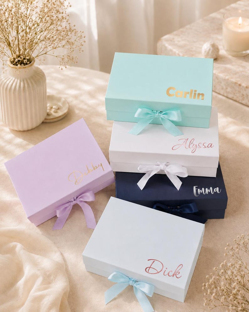 May include: A collection of personalized gift boxes in various pastel colors, including light blue, lavender, white, and navy. Each box is adorned with a matching ribbon and features a name in gold or rose gold script. The boxes are arranged on a neutral surface.
