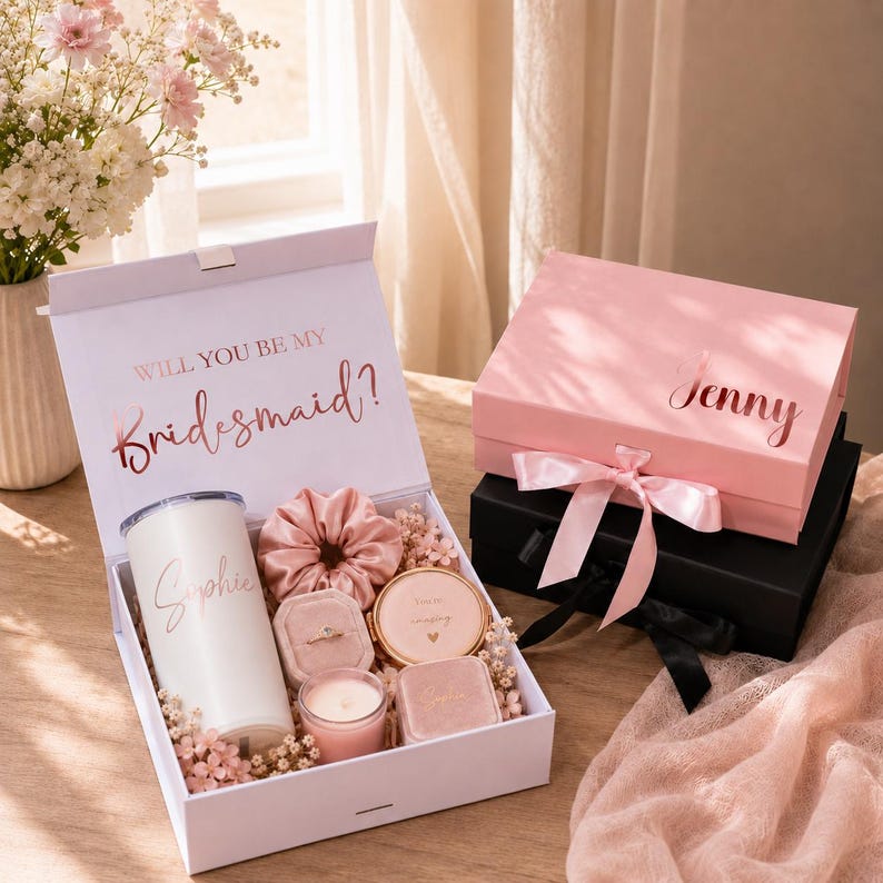 May include: A white gift box with the text "WILL YOU BE MY Bridesmaid?" contains a tumbler, scrunchie, ring box, compact mirror, and candle. A pink box with "Jenny" on top is tied with a pink ribbon. A black box is beneath the pink box.