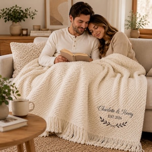 May include: A cream-colored textured throw blanket with fringe edges, featuring embroidered black text that reads Charlotte & Henry EST.2026 above a decorative leaf branch design. The blanket is draped over a sofa, covering two people.