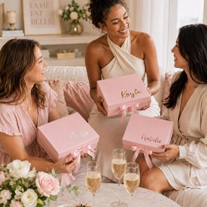 May include: Three women are gathered, each holding a pink gift box with a name on it. The boxes have matching pink ribbons. Champagne flutes and a plate of macarons and strawberries sit on a table. A framed print reads "La Vie Est Belle."