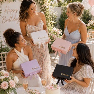 May include: Four women holding personalized gift boxes in white, pink, lavender, and black. Each box has a name in gold script and a matching ribbon. The scene is set outdoors with floral decorations and a "Happy Birthday" sign.