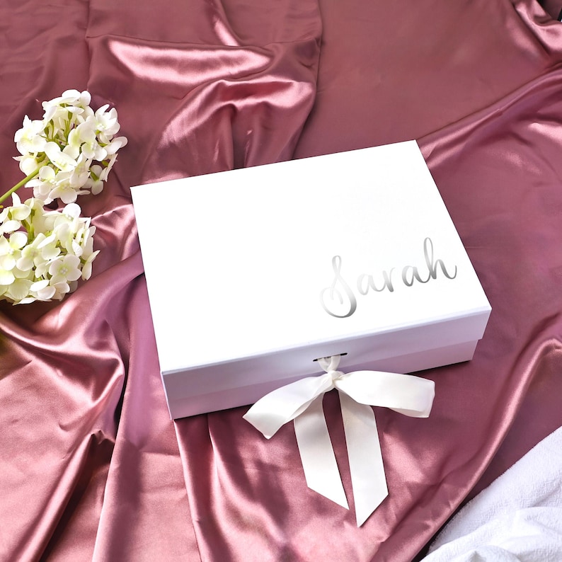 May include: A white gift box with the name "Sarah" in silver script on the lid. The box is tied with a white satin ribbon and sits on a pink satin surface, with white flowers in the background.