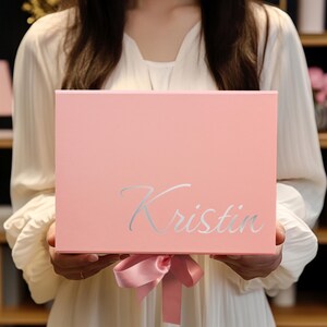 May include: A light pink gift box with the name "Kristin" in silver script. The box is tied with a matching pink satin ribbon. The box is held by a person wearing a white dress.