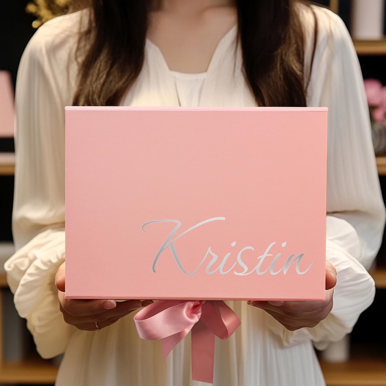 May include: A light pink gift box with the name "Kristin" in silver script. The box is tied with a matching pink satin ribbon. The box is held by a person wearing a white dress.