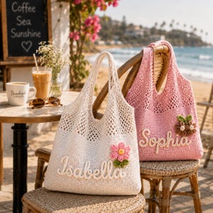 May include: Two crochet tote bags with open-knit tops and solid-knit bottoms. The white bag features the name Isabella in white script with a pink flower appliqué. The pink bag features the name Sophia in white script with a brown flower appliqué.