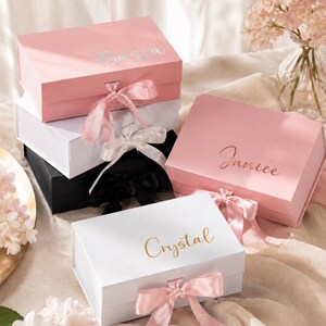 May include: A collection of personalized gift boxes in various colors, including pink, white, and black. Each box features a name in elegant script, such as "Crystal" and "Janice", and is adorned with a matching satin ribbon. The boxes are arranged on a soft, neutral-toned surface.