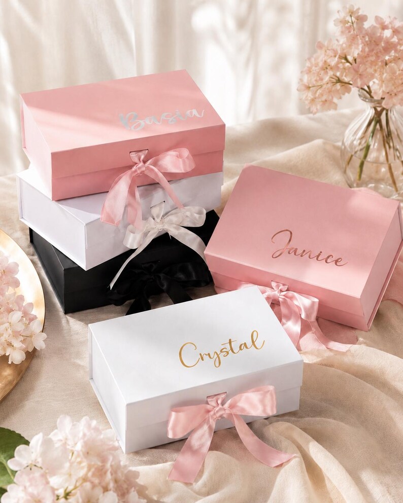 May include: A collection of personalized gift boxes in various colors, including pink, white, and black. Each box features a name in elegant script, such as "Crystal" and "Janice", and is adorned with a matching satin ribbon. The boxes are arranged on a soft, neutral-toned surface.