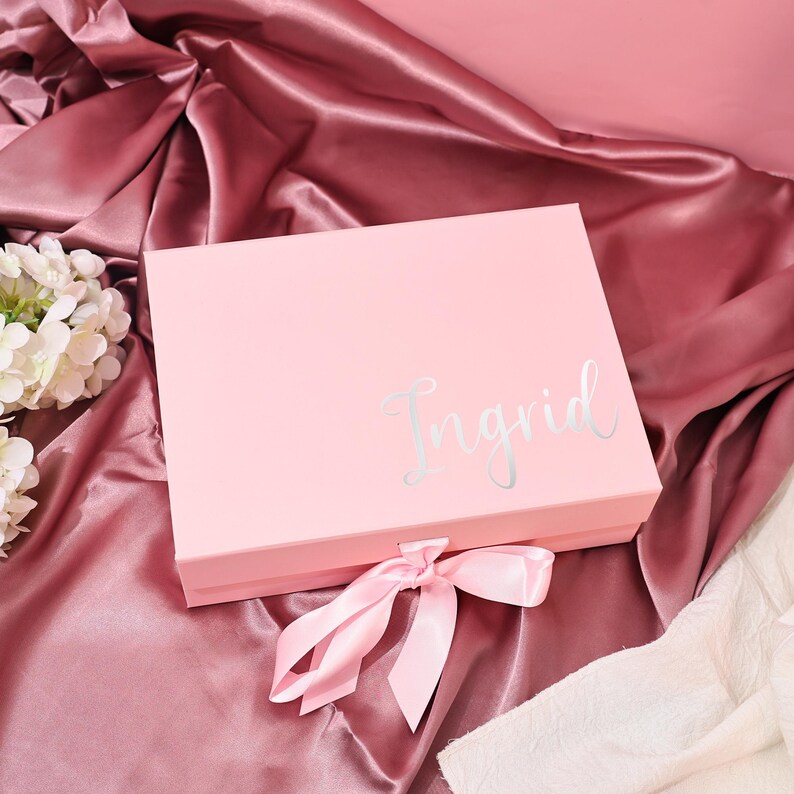 May include: A light pink gift box with the name "Ingrid" in silver script. The box is tied with a matching pink satin ribbon. The box is on a pink satin fabric background.