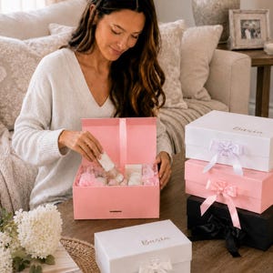 May include: A woman opening a pink gift box filled with white items. Several other gift boxes are stacked nearby, in white, pink, and black, each with a ribbon. The boxes have the word "Basta" printed on them.