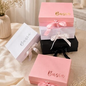 May include: A collection of gift boxes in various colors, including pink, white, and black. Each box is adorned with a satin ribbon and the name "Basia" in a metallic font. The boxes are arranged on a neutral surface, with decorative flowers in the background.