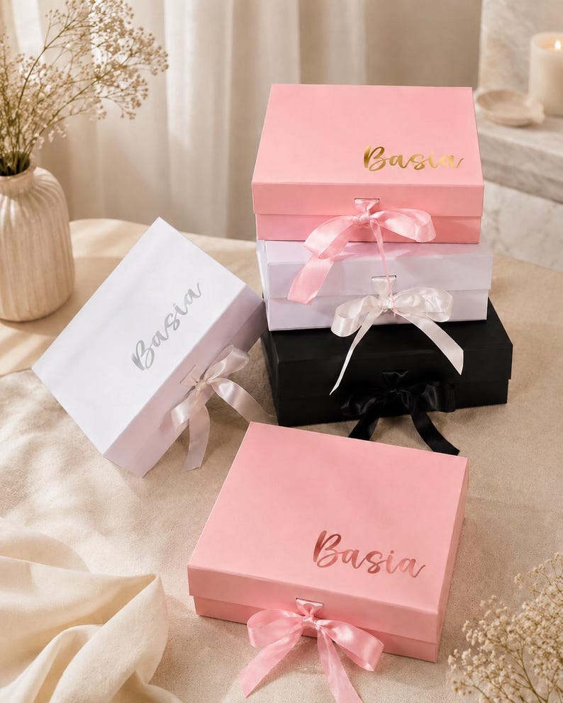 May include: A collection of gift boxes in various colors, including pink, white, and black. Each box is adorned with a satin ribbon and the name "Basia" in a metallic font. The boxes are arranged on a neutral surface, with decorative flowers in the background.