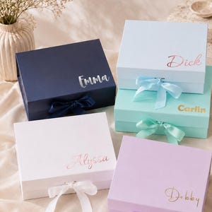 May include: Four personalized gift boxes in navy, white, light blue, and lavender. Each box has a matching ribbon and a name in a metallic script: Emma, Dick, Alyssa, and Debby. The boxes are closed and ready for gifting.