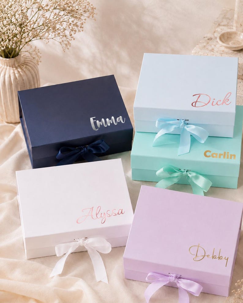 May include: Four personalized gift boxes in navy, white, light blue, and lavender. Each box has a matching ribbon and a name in a metallic script: Emma, Dick, Alyssa, and Debby. The boxes are closed and ready for gifting.