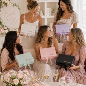 May include: A group of women holding gift boxes in various pastel colors, each with a name on top. The boxes have matching ribbons. A sign that says "Bride to Be" is in the background. Champagne glasses and a cake are on the table.
