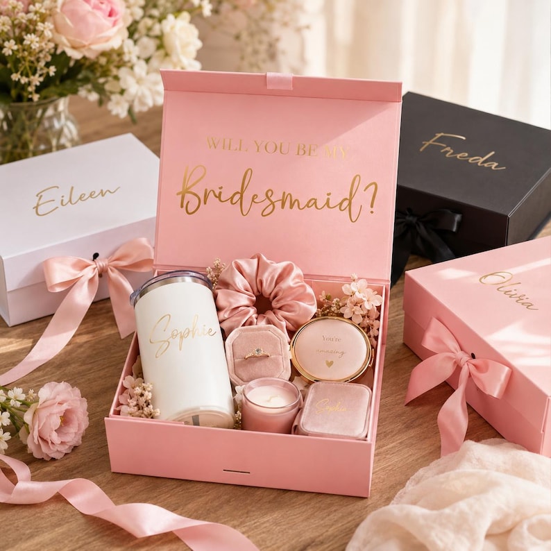 May include: A pink gift box with the text "Will You Be My Bridesmaid?" in gold script. Inside, a white tumbler, scrunchie, ring box, compact mirror, and candle. Other boxes with names are also visible.