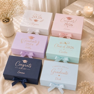 May include: Six graduation gift boxes in pastel shades of white, pink, lilac, mint, and navy. Each box is adorned with a ribbon and text like "Class of 2026" and a name. The boxes are decorated with graduation-themed graphics.