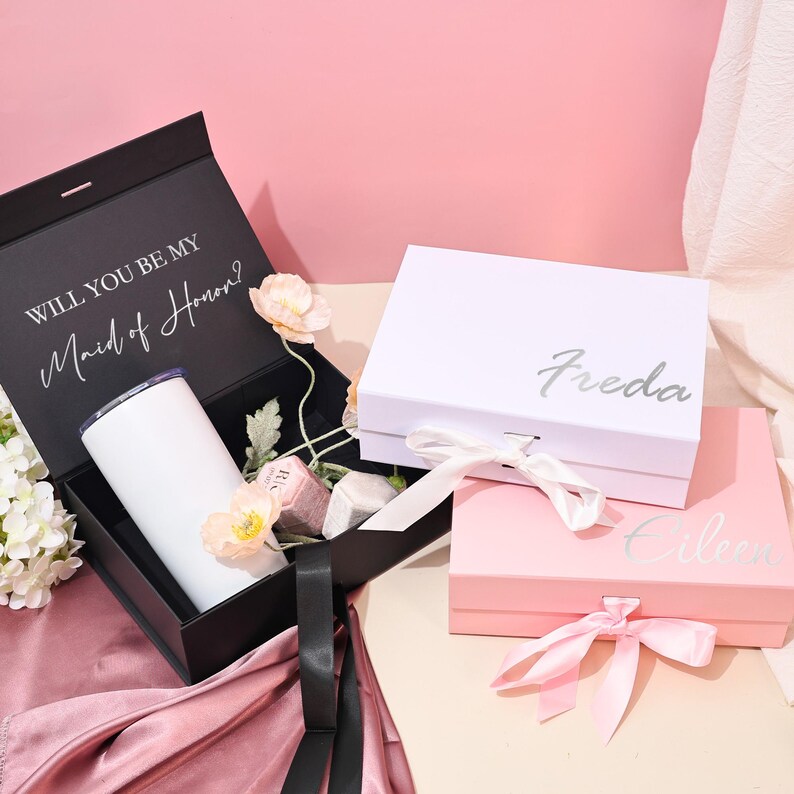 May include: A black gift box with the text "WILL YOU BE MY Maid of Honor?" and a white tumbler. Two other boxes are visible, one white with "Freda" and one pink with "Eileen" written on them. All boxes have ribbons.