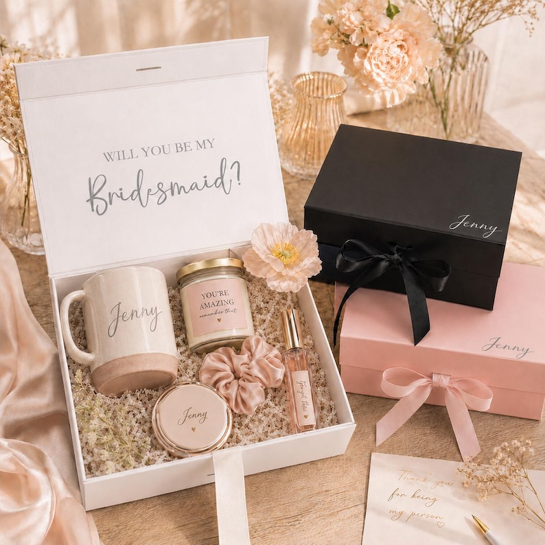 May include: A white gift box with the text "Will You Be My Bridesmaid?" contains a mug, candle, scrunchie, compact mirror, and perfume. A black box and a pink box are also visible, each with a name on them. The boxes are tied with ribbons.
