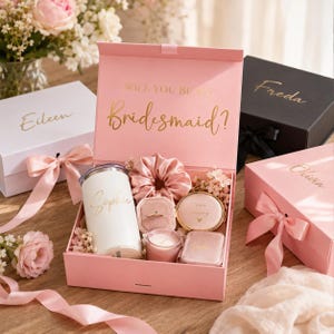 May include: A pink gift box with gold lettering reading "Will You Be My Bridesmaid?" contains a white tumbler, scrunchie, ring box, compact mirror, and candle. Other boxes with names are also visible.