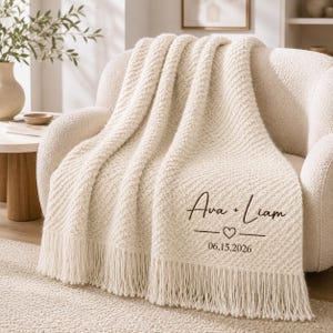 May include: A cream-coloured knit throw blanket with a textured chevron pattern and long fringe edges. The blanket features dark brown printed text that reads Ava + Liam, a small heart icon, and the date 06.15.2026 centred on the fabric.