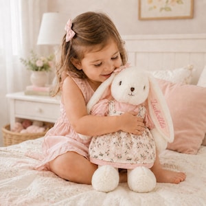 Könnte beinhalten: Ein cremefarbenes Plüschhasenspielzeug mit langen Ohren, das ein rosa Kleid mit Blumenmuster trägt. Der Name Isabella ist mit rosa Faden auf eines der Ohren des Hasen gestickt. Das Spielzeug hat eine weiche Textur und eine kleine rosa Schleife.