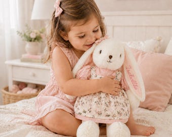 May include: A plush cream-colored rabbit toy with long ears, wearing a pink dress with a floral pattern. The name Isabella is embroidered in pink thread on one of the rabbit's ears. The toy has a soft texture and a small pink bow on its head.