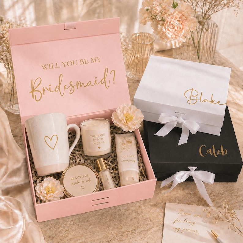May include: A pink gift box with the text "WILL YOU BE MY Bridesmaid?" in gold lettering. Inside, a mug with a heart, a candle, and other beauty products. Two other gift boxes, one white and one black, are also visible.