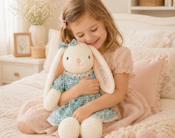 May include: A cream-colored plush rabbit toy with long ears, wearing a teal floral dress with a white lace collar and a matching teal bow on its head. The name Olivia is embroidered in teal thread on one of the rabbit's long, soft ears.