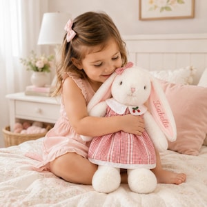 May include: A plush white rabbit toy with long ears, wearing a red and white gingham dress with a white collar featuring a small cherry embroidery. The name Isabella is embroidered in pink thread on one ear, and a small red gingham bow is on its head.