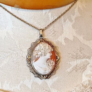 Könnte beinhalten: Eine silberne Kettenkette mit einem Kameenanhänger. Die Kamee ist eine ovale Muschel mit einem geschnitzten Bild eines Frauenprofils. Die Kamee ist in einen goldfarbenen Filigranrahmen gefasst.