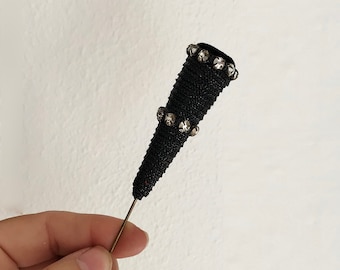 May include: A black beaded pin with a pointed end and silver rhinestones. The pin is held in a hand.