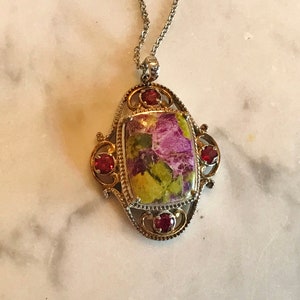 May include: A silver chain necklace with a pendant featuring a green and purple stone set in a gold-toned filigree frame with red gemstone accents.