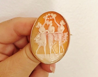 May include: A cameo brooch featuring three women in a delicate dance, carved from shell with a gold tone finish. The women are holding a wreath of flowers above their heads.