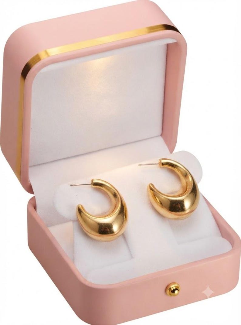 May include: A pair of polished gold-tone hoop earrings with a thick, rounded profile. The earrings are displayed inside an open, light pink rectangular jewelry box featuring a white interior lining and a small gold-colored latch on the front.