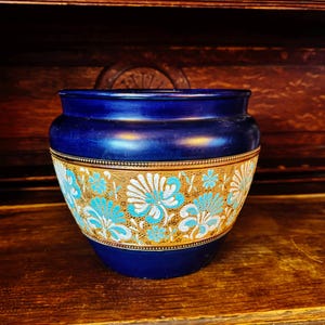 May include: A cobalt blue ceramic pot with a gold band featuring a floral design in white and turquoise. The pot has a rounded shape and sits on a wooden surface, showcasing a vintage aesthetic.
