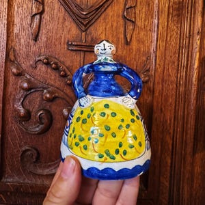 Op de afbeelding: Handbeschilderde keramische figuur van een vrouw in een blauwe top en gele schort met groene stippen. Het beeldje heeft een witte kop en een blauw-wit patroon rok. Gehouden voor een houten achtergrond.