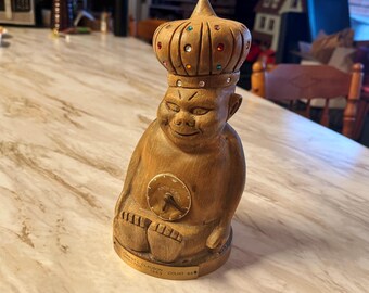 May include: A wooden figurine of a gnome wearing a crown with jewels. The gnome is sitting with its legs crossed and holding a small clock in its hands. The base of the figurine has the text "NORTH STAR SALOON COUNT 845".