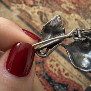 Puede incluir: Un broche de metal plateado con un dise&ntilde;o detallado de hojas y un mecanismo de alfiler visible en el reverso. La superficie met&aacute;lica muestra un acabado texturizado y envejecido con venas intrincadas en las hojas y un alfiler fino y curvado.