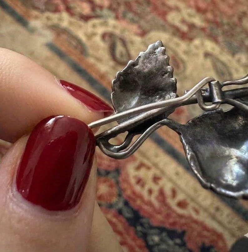 Puede incluir: Un broche de metal plateado con un dise&ntilde;o detallado de hojas y un mecanismo de alfiler visible en el reverso. La superficie met&aacute;lica muestra un acabado texturizado y envejecido con venas intrincadas en las hojas y un alfiler fino y curvado.