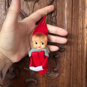 Puede incluir: Un adorno de elfo navideño vintage. El elfo tiene una cara de plástico con ojos azules, un gorro de fieltro rojo y un atuendo de fieltro rojo con ribete blanco. El adorno está sostenido por una mano.