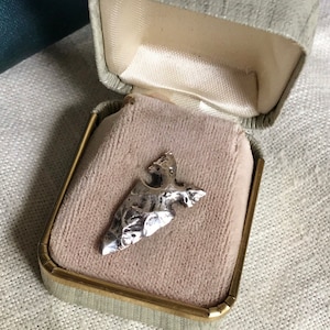 Puede incluir: Un colgante de plata con forma de punta de flecha descansa dentro de un joyero vintage. El colgante tiene una superficie texturizada y se muestra sobre un forro de terciopelo beige rosado. La caja es verde claro con un borde dorado.