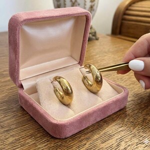 May include: A pair of thick, polished gold hoop earrings with a smooth, rounded surface. The earrings are displayed inside an open, square-shaped jewelry box covered in soft, dusty pink velvet with a light beige satin interior lining.