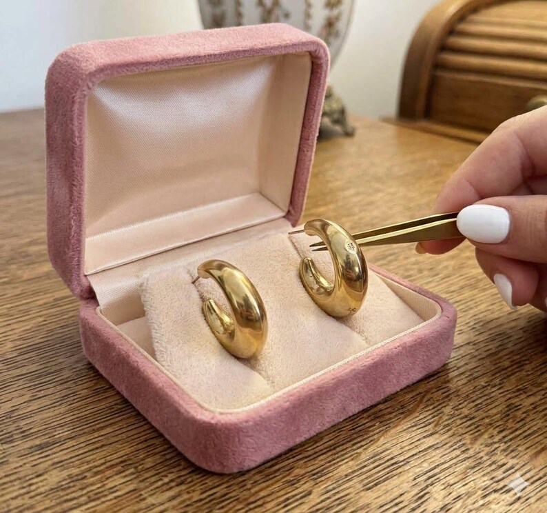 May include: A pair of thick, polished gold hoop earrings with a smooth, rounded surface. The earrings are displayed inside an open, square-shaped jewelry box covered in soft, dusty pink velvet with a light beige satin interior lining.