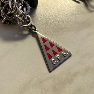 May include: A silver-toned charm with a triangular pendant featuring red triangles and the letters "C.F.G." attached to a chain. The charm is likely part of a bracelet or necklace.