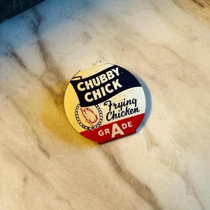 May include: A round vintage sign with the text "CHUBBY CHICK Frying Chicken Grade A". The sign has a white background with a blue and red border. A gold chain surrounds a red chicken illustration.